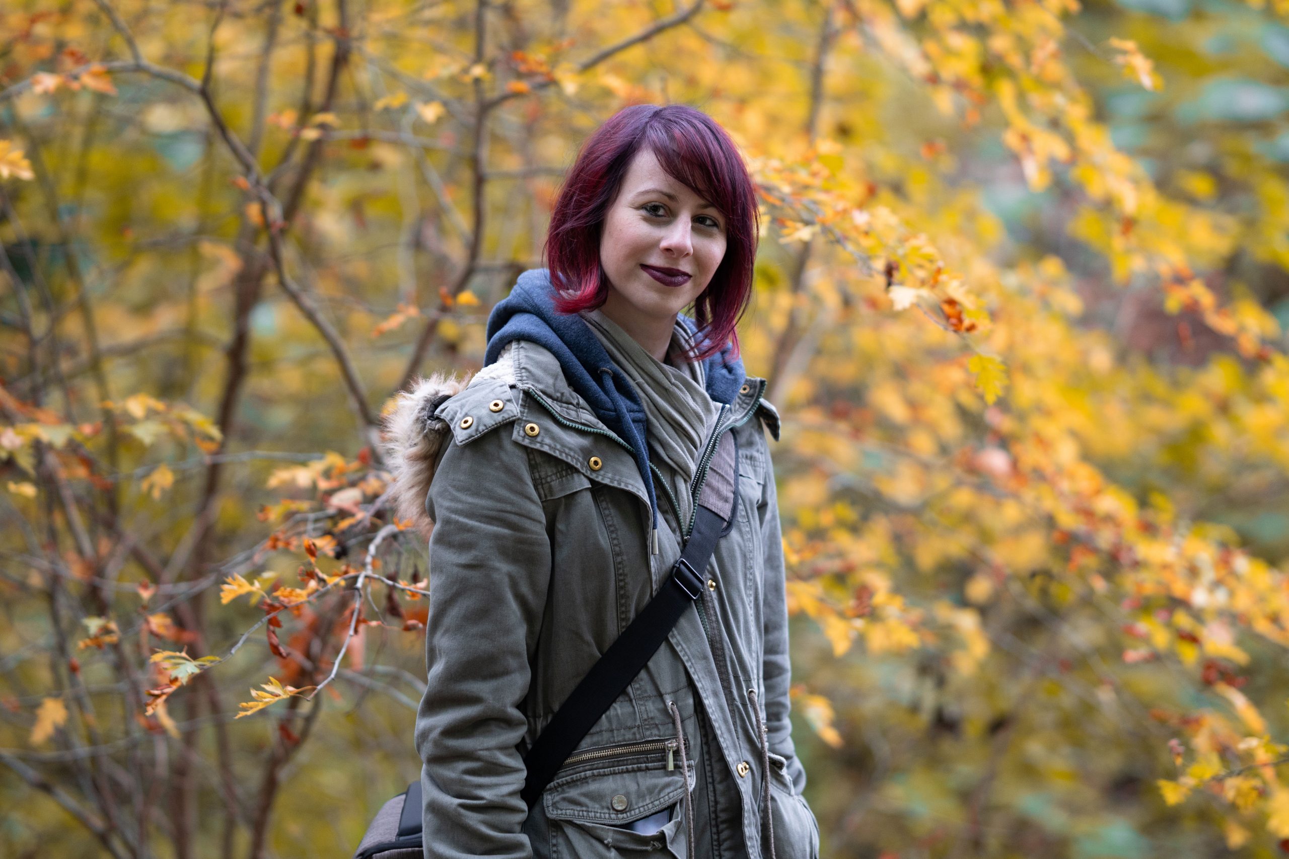 Profilbild: Frau mit roten Haaren, steht vor Herbstbäume, schaut von der Seite in die Kamera